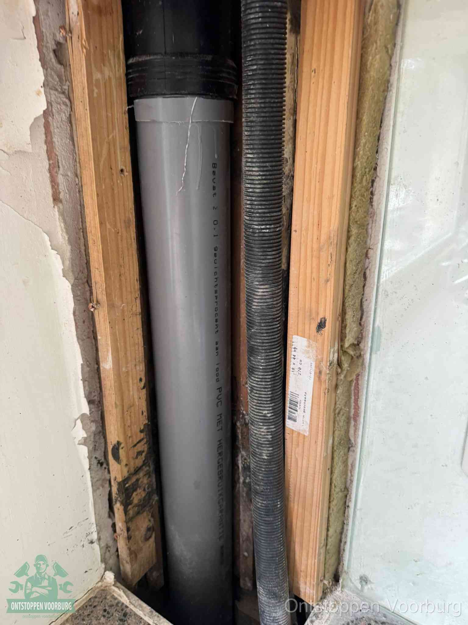 Een grijze PVC-afvoerbuis met een flexibele ribbelslang in een open wandconstructie, een verwaarloosde afvoer die te lang is blijven zitten in Voorburg
