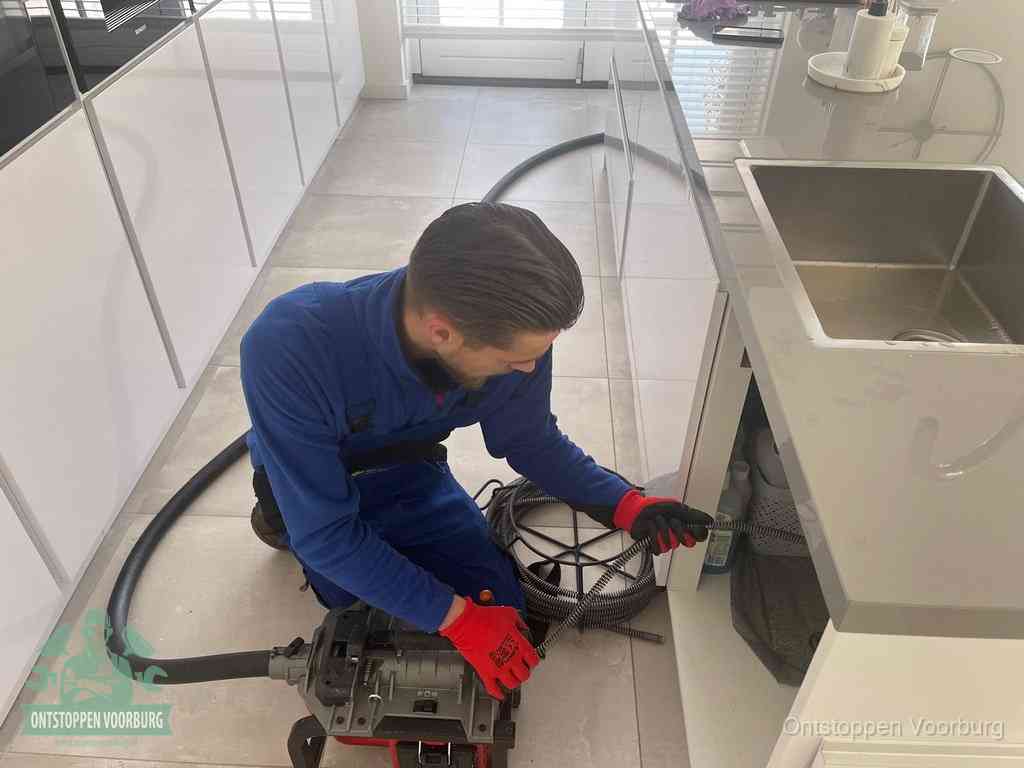 Een monteur ontstopt de keukenafvoer onder de spoelbak met een elektrische veermachine in Voorburg
