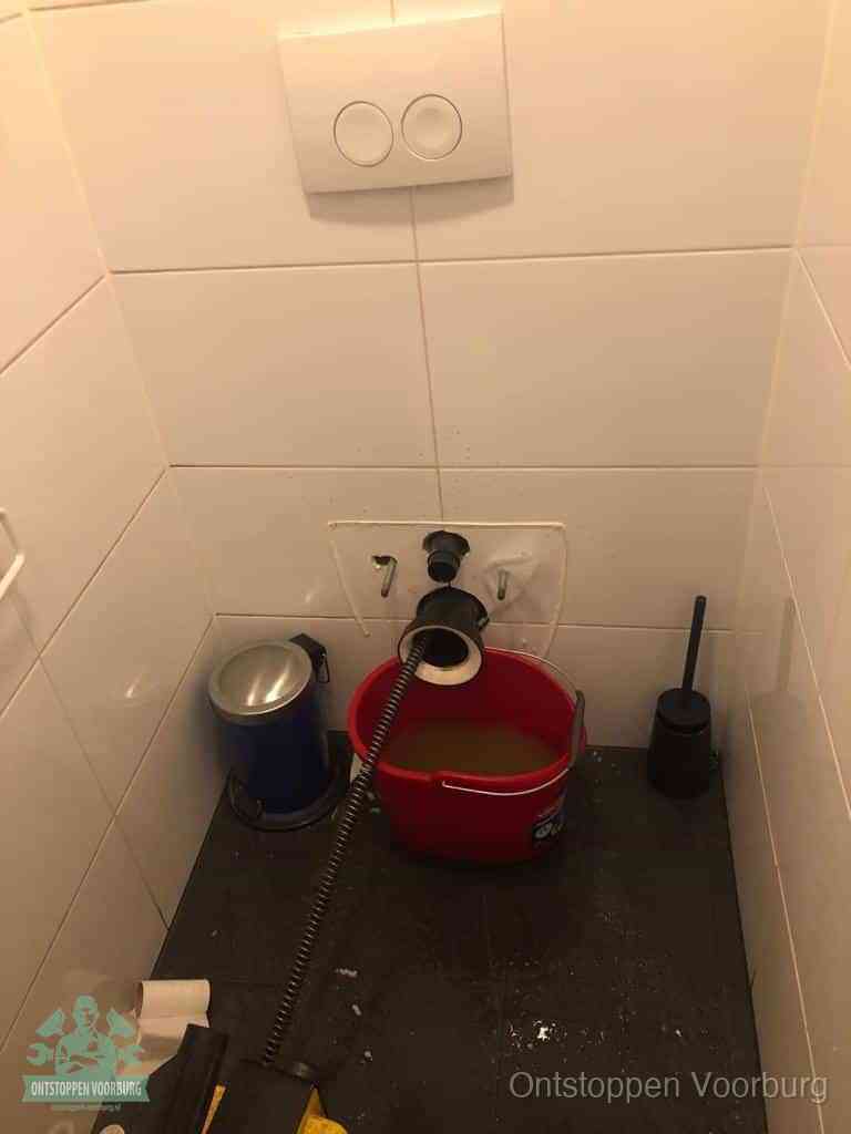 Een ontstopte wand-wc met verwijderde toiletpot waar een ontstoppingsveer in de wanduitgang verdwijnt, het achterhalen waar de verstopping zit in Voorburg