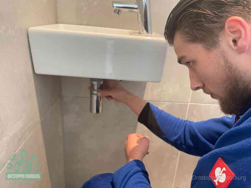 Een monteur draait met de hand de zwanenhalssifon onder een fonteinwasbak in de toiletruimte los om de bron van rioollucht in Voorburg te vinden