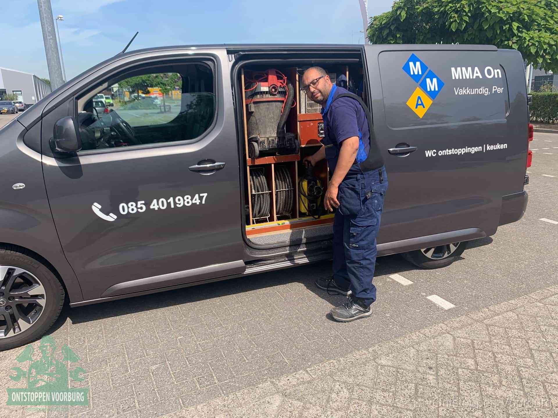 Een monteur bij de open schuifdeur van zijn bedrijfsbus op een parkeerplaats, klaar om de oorzaak van terugkerende afvoerverstoppingen aan te pakken in Voorburg