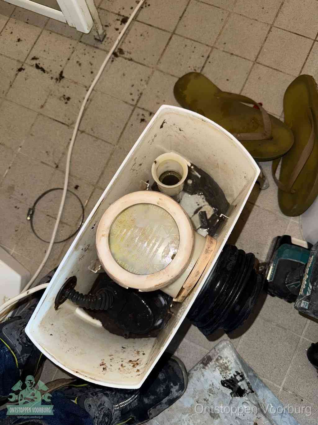 Een gedemonteerde toiletpot op de vloer met ontstoppingsveer en gereedschap ernaast, het verwijderen van een vastzittend voorwerp uit de wc in Voorburg