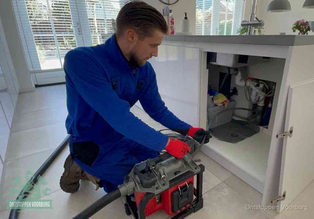 Een monteur voert een professionele elektrische ontstoppingsmachine in de afvoer onder de spoelbak tijdens het gootsteen ontstoppen in Voorburg